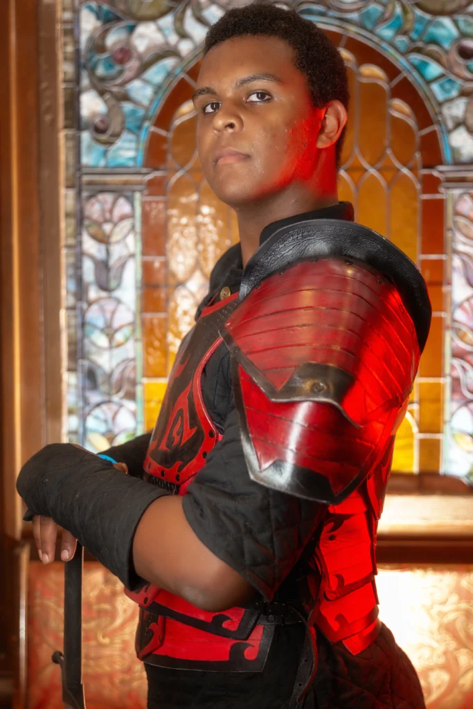 Justin Cale II wearing his signature black and red armor posing against a glass mosiac arch background.