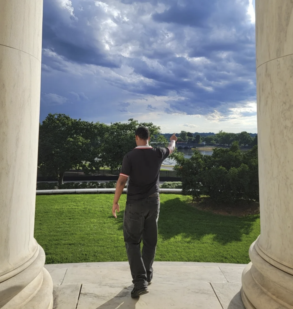 Justin Cale II stands between two marble columns in Washington, D.C., facing a grassy park and river while pointing toward a dramatic cloudy sky.