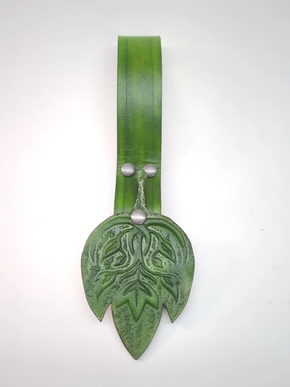 A close up of the front of a green skirt hike against a white background. Displays the skirt hike's green strap and embossed fantasy leaf design on the emblem.