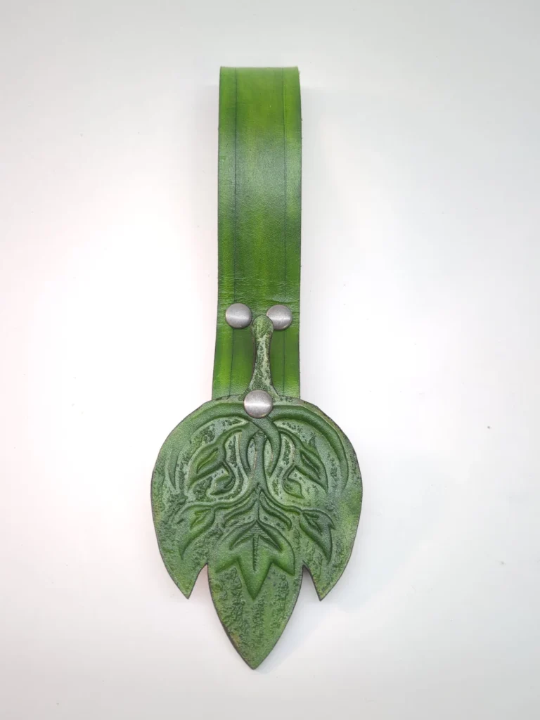A close up of the front of a green skirt hike against a white background. Displays the skirt hike's green strap and embossed fantasy leaf design on the emblem.
